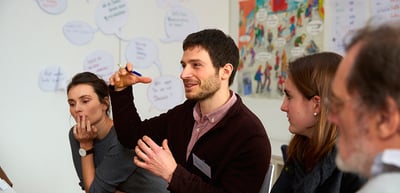A young man raising his hand while speaking in a discussion group in front of a wall with notes