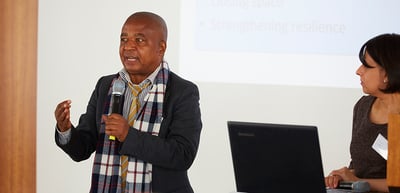 Man in a checked scarf speaking into a microphone next to a woman at a laptop