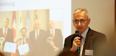 Man with glasses speaking into a microphone in front of a projected photo of award winners