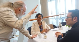 Older man with a white beard leaning over a table to talk with a woman and a man