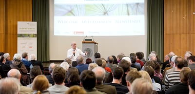 Speaker with a microphone in front of a full lecture hall, the screen reads "Welcome · Willkommen · Bienvienue" and a banner reads "On the way to Rio+20"