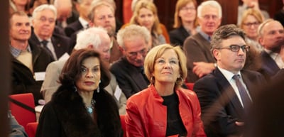 Audience in the front rows of a hall, a woman in a red jacket looking up