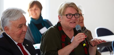 Woman with glasses speaking into a microphone next to an older man
