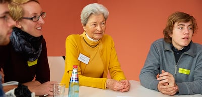 A laureate in a yellow sweater sitting between two young participants at a round table