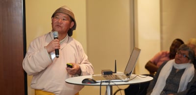 Man in a traditional hat and a light robe speaking into a microphone next to a laptop