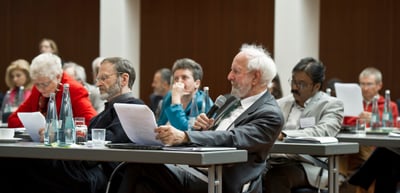 Older man with a white beard speaking into a microphone at a conference table