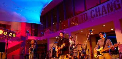 Band performing on a stage lit in blue and red, the wall lettering reads "Power to Change"