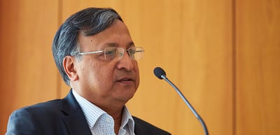 Man with glasses speaking into a microphone in front of a wooden wall