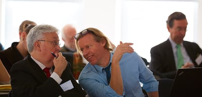 Two men at a conference table, one leaning towards the other and smiling
