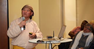 Man in a traditional hat and a light robe speaking into a microphone next to a laptop