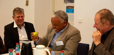 A laureate holding an apple and laughing with two other laureates at a round table
