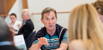 Man in a striped polo shirt explaining something with both hands during a discussion